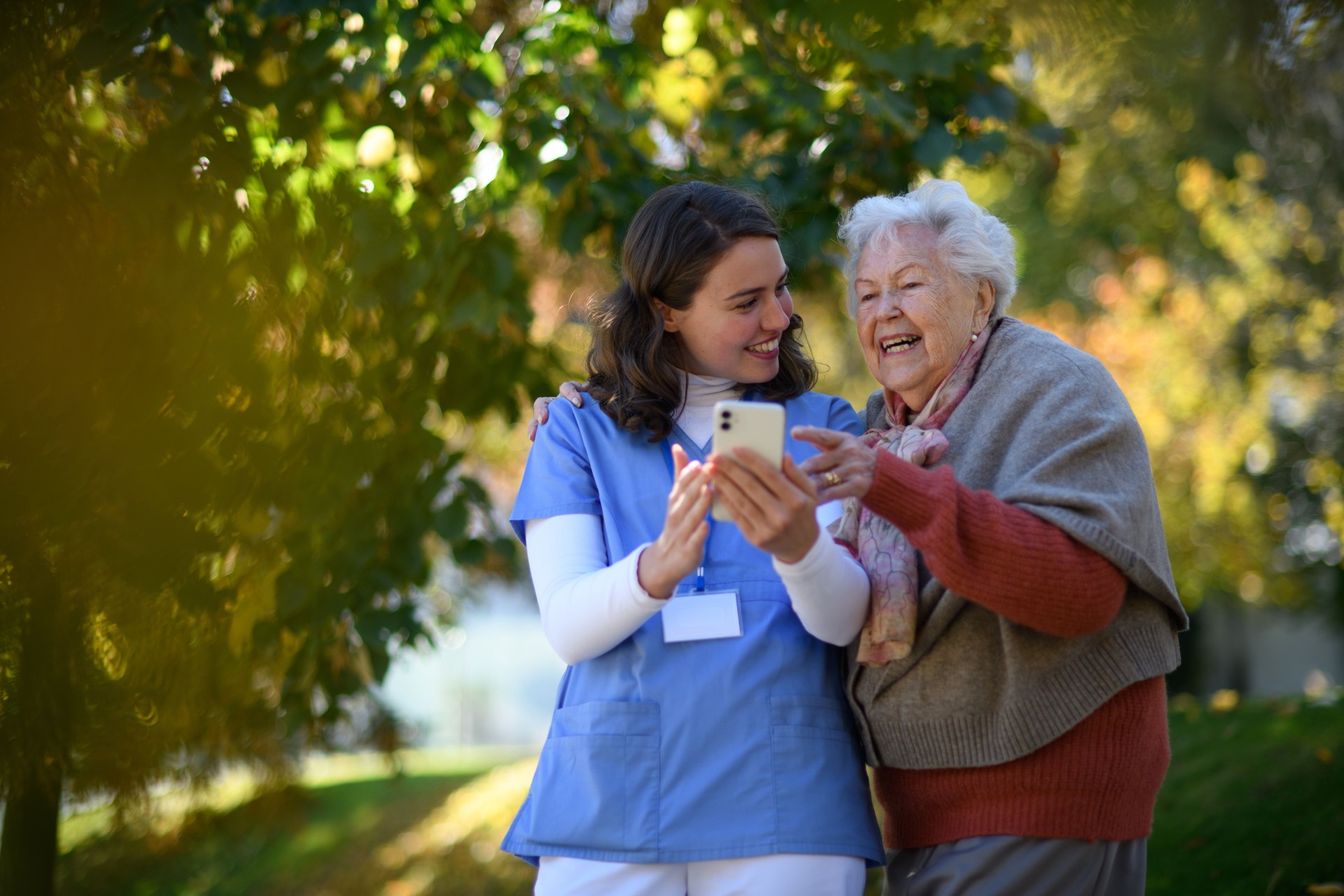 Portrait of nurse and elderly woman on walk in the park during warm autumn day. Young caregiver looking through photos of senior patient's family on a phone.